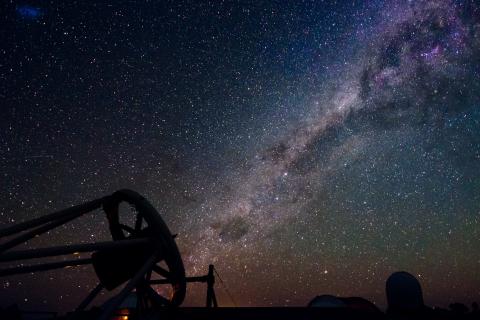 In the lower left corner, a telescope is pointing at the night sky towards the upper right. The band of the Milky Way, a bright and dusty thick line, runs from the telescope to the top right corner. Many white dots of stars are across the sky, varying in size. The sky is mostly a blue-black, but is a yellow-red towards the bottom horizon. There is a silhouette of a telescope dome towards the lower right of the image.