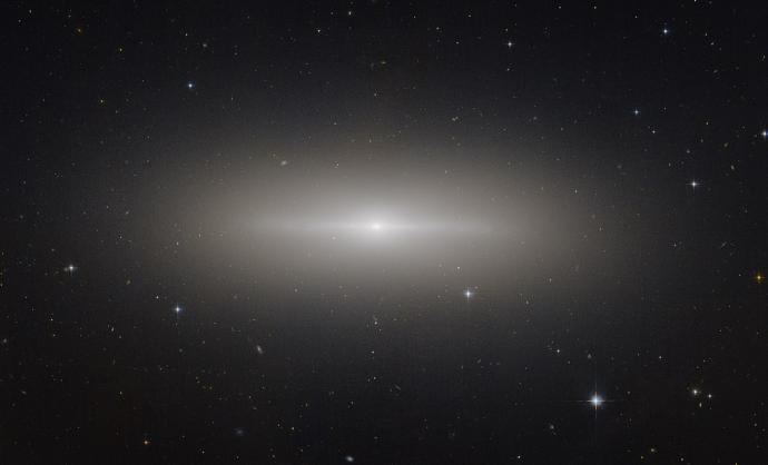 Small white dots of stars against a black background, some slightly larger than others. In the middle, a white sphere, roughly similar in size to some of the larger stars, glows veery brightly. There is a cloudy, disk-like structure around the sphere, almost flattened along the horizontal. A soft, diffuse light glows around it