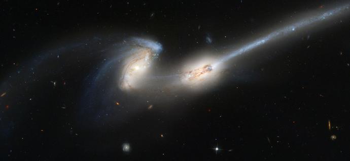 Two galaxies against a black background, in which some small dots of stars and other smaller galaxies are visible in various colours. The pair of galaxies in the middle have bright, yellow-white centers, giving off a glow. The left galaxy has spiral arms, though the lower one is pulled towards and into the galaxy on the right. From this galaxy, a bright blue-grey, cloudy streak travels towards the upper right corner of the image.