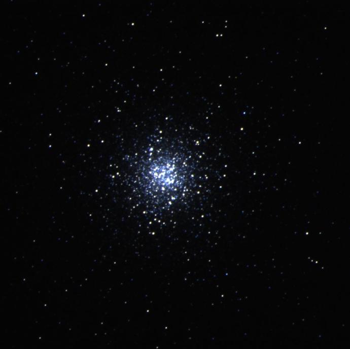 An image of a star cluster against a black background. Small dots of stars are mostly concentrated in the centre, many of them white in colour but some of them having a blue hue to them. Further out from the middle, a few more stars are scattered around, but they are fewer in number and seem more reddish compared to the others.