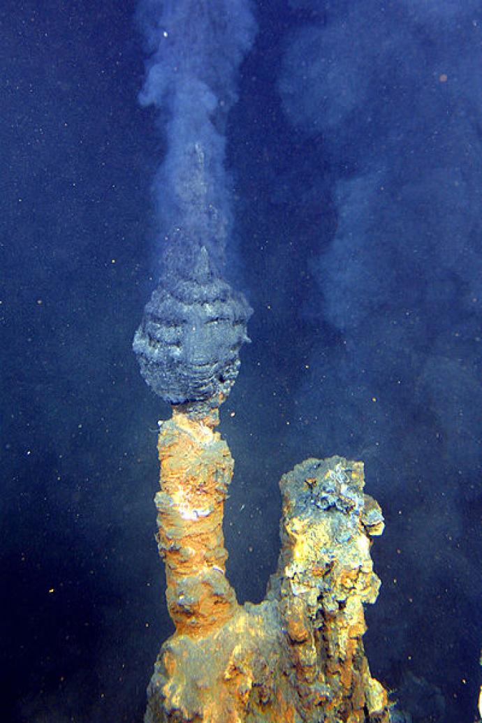 A photograph of a black smoker, an underwater hydrothermal vent located on the ocean floor. The structure releases dark clouds of mineral-rich water, which are visible as it rises from the chimney-like formation, creating a striking contrast against the deep blue background of the ocean