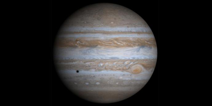 Image of Jupiter showcasing its characteristic bands of clouds, with the Great Red Spot visible on the right. A shadow of one of its moons can be seen as a dark spot on the planet's surface