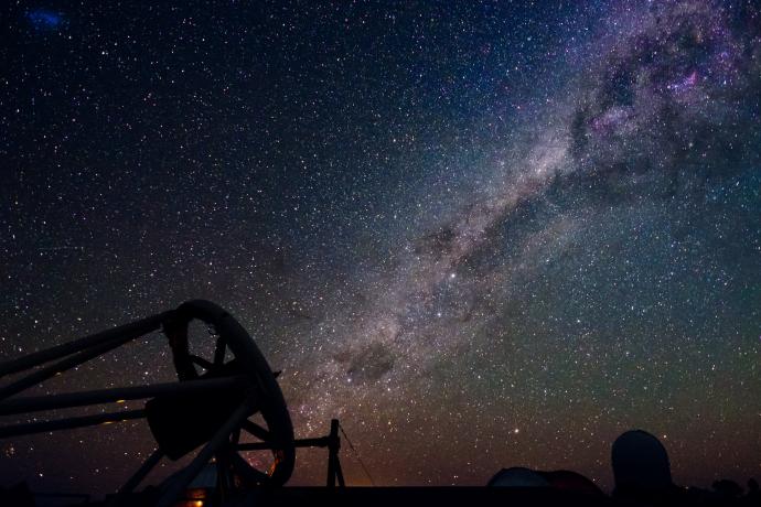 In the lower left corner, a telescope is pointing at the night sky towards the upper right. The band of the Milky Way, a bright and dusty thick line, runs from the telescope to the top right corner. Many white dots of stars are across the sky, varying in size. The sky is mostly a blue-black, but is a yellow-red towards the bottom horizon. There is a silhouette of a telescope dome towards the lower right of the image.