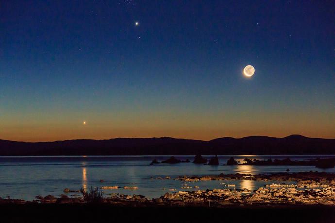 A serene twilight landscape showing a crescent moon above a calm body of water, with Venus and Jupiter shining brightly in the sky. The reflection of the moon can be seen on the water, with silhouettes of distant mountains and rocky terrain in the foreground